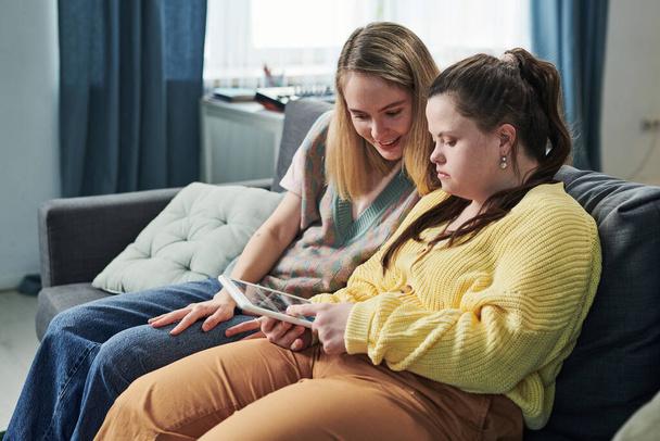Moderne jonge vrouw met het syndroom van Down tijd doorbrengen met haar zus thuis kijken naar inhoud in sociaal net op digitale tablet - Foto, afbeelding