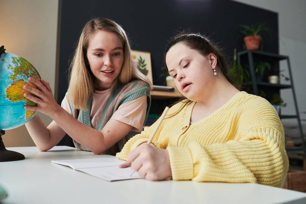Giovane insegnante di sesso femminile seduta a tavola accanto al suo studente con sindrome di Down che aiuta con il compito durante la lezione di geografia - Foto, immagini