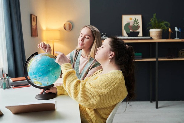 Jong meisje met Down syndroom zitten aan het bureau naast haar leraar draaien globe en praten over landen en culturen - Foto, afbeelding