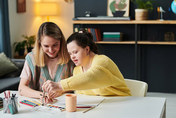 Mädchen mit Down-Syndrom wählt Aquarellfarbe aus Palette mit Pinsel während des individuellen Kunstunterrichts mit junger Lehrerin zu Hause - Foto, Bild