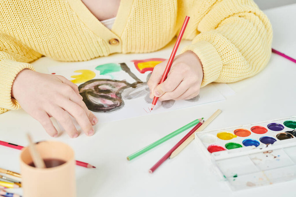 Высокоугольный средний крупный план неузнаваемой девушки, сидящей за столом и рисующей красочную картину на бумаге акварельными красками и карандашами - Фото, изображение