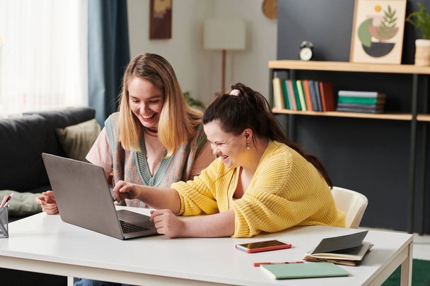 Молодая белая женщина и ее университетская подружка с синдромом Дауна сидят за столом дома и смеются над чем-то смешным во время выполнения домашнего задания - Фото, изображение