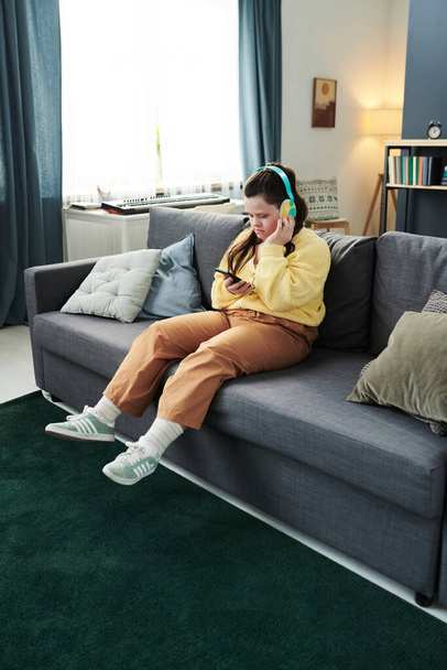 Vertical largo tiro de mujer joven con síndrome de Down con auriculares sentados relajados en el sofá en la sala de estar encender la música con el teléfono inteligente - Foto, imagen
