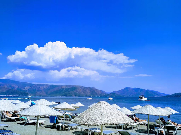 a scene with the sea, mountains, blue sky, and straw umbrellas providing shade from the sun - Photo, Image