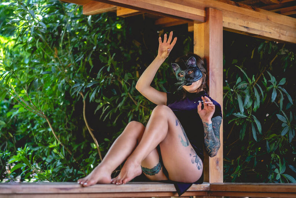 Egzotik baştan çıkarma: dövmeli seksi esmer kadın mavi Çinli elbiseli, tahta bir çadırda kedi maskesi takan, ormanla çevrili. - Fotoğraf, Görsel