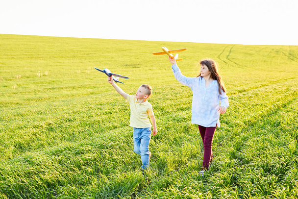 Running boy and girl holding two yellow and blue airplanes toy in the field during summer sunny day - Фото, зображення