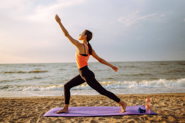 Modern young woman doing yoga on the beach at sunrise. Sports woman doing stretching outdoors. The concept of an active lifestyle, sports training, yoga or fitness. - Photo, Image