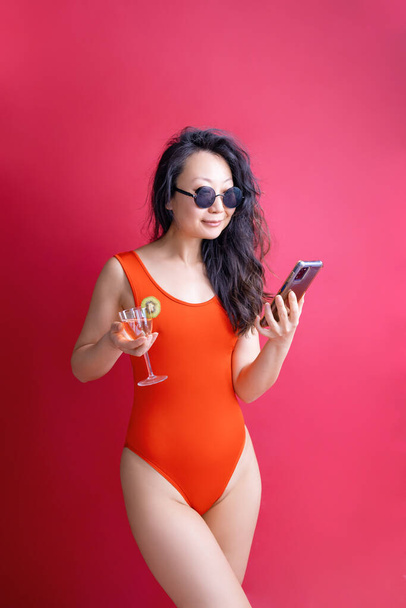 Jeune femme d'origine asiatique en maillot de bain rouge tenir cocktail téléphone portable isolé sur fond rouge. Vacances d'été mer repos soleil bronzage concept. - Photo, image