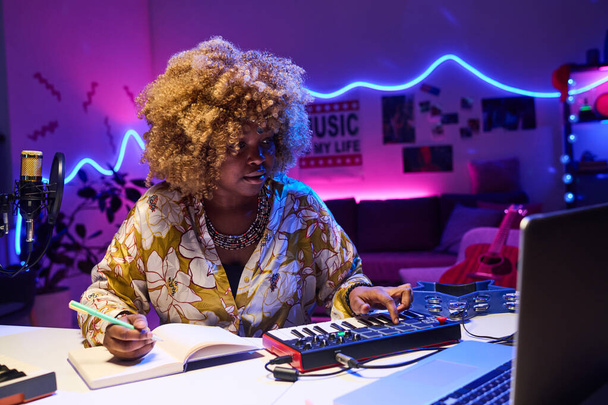 Horizontal medium portrait of stylish young African American woman sitting at desk at home composing song using synthesizer and laptop - Фото, зображення