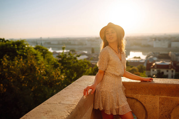 Portrait of a happy woman tourist in a hat at dawn enjoys the cityscape. A young woman is traveling in Europe. The concept of an active lifestyle, tourism or vacation. - Foto, Imagen