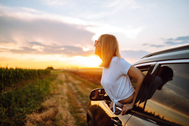 Lifestyle! Happy woman enjoys sunset views from the car window. Young tourist woman rests and leans out of the car window, enjoys the trip. Travel concept, vacation. Towards adventure. - Фото, изображение