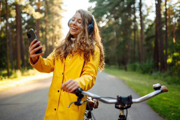 Ευτυχισμένη νεαρή γυναίκα με ακουστικά και smartphone βόλτες με ποδήλατο σε ένα ηλιόλουστο πάρκο ακούγοντας μουσική, podcast. Χαλάρωσε, έννοια της φύσης. Τρόπος ζωής. - Φωτογραφία, εικόνα