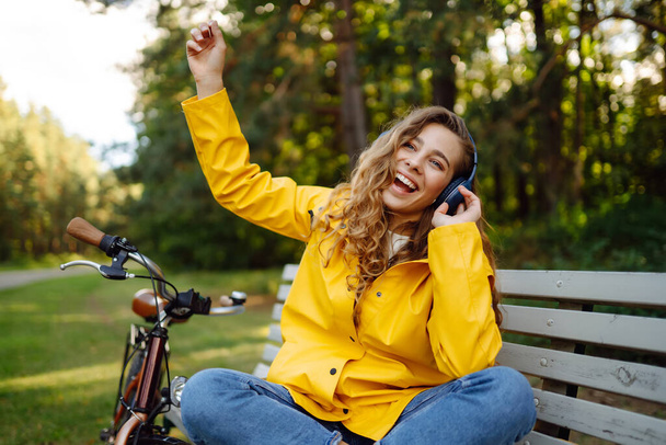 Happy young woman with headphones and smartphone rides a bicycle in a sunny park listening to music, podcast. Relax, nature concept. Lifestyle. - Фото, зображення