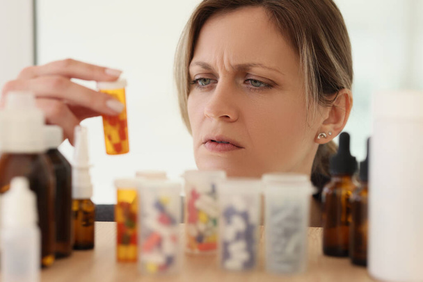Focused woman reads instruction on plastic bottle with pills. Female person looks for medication to relieve acute headache after work in office - Фото, изображение