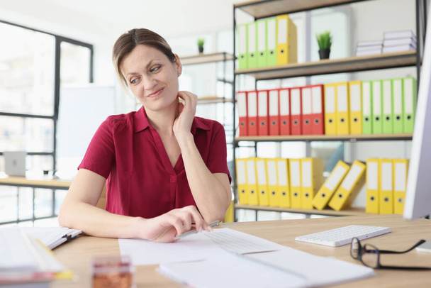 Young woman sits at wooden table looking aside and pursing lips. Exhausted secretary feels lazy to continue accounting paperwork in office - Foto, Imagen