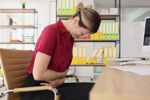 Woman feels acute stomach pain at workplace in office. Young female secretary tries to handle abdominal spasms sitting at table in office - Foto, imagen