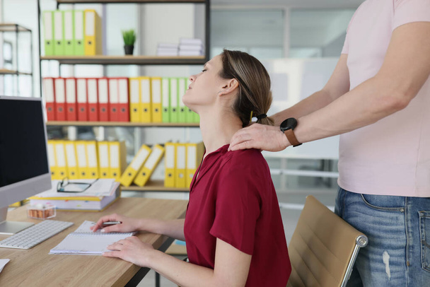 Male colleague massages neck of tired woman with strong hands. Female employee in red blouse sits at wooden table in office enjoying massage - Foto, afbeelding