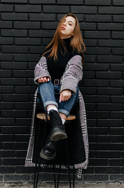 uma menina em um casaco cinza senta-se em uma cadeira perto de uma parede preta - Foto, Imagem