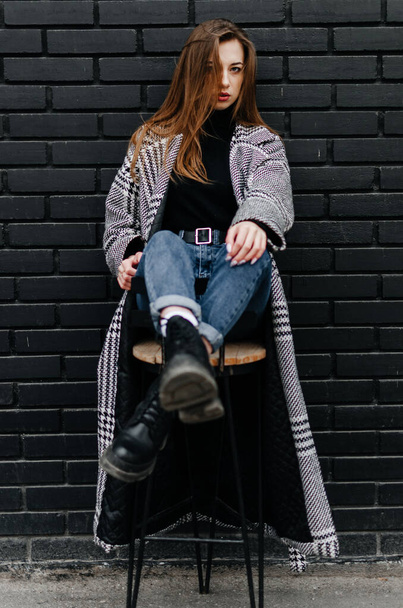 Девушка в сером пальто сидит на стуле возле черной стены - Фото, изображение