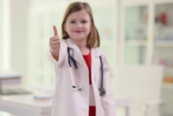 Schattig klein meisje in uniform van de dokter toont duim omhoog gebaar. Wazig kind met stethoscoop staat in het ziekenhuis kantoor op zoek in camera met glimlach - Foto, afbeelding