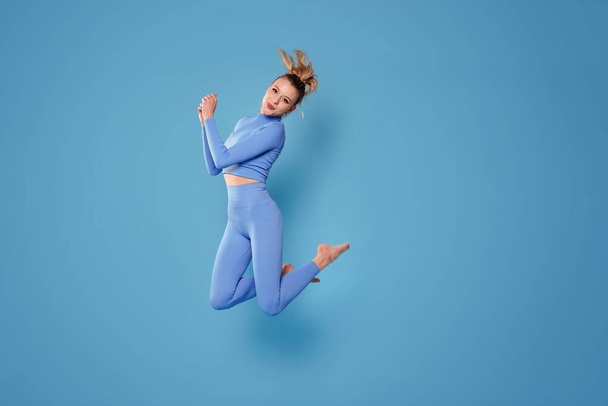 Szczęśliwa, wysportowana kobieta w modnej odzieży sportowej skacząca na niebieskim tle studia. Sport i zdrowy styl życia. Dużo miejsca na kopie.. - Zdjęcie, obraz