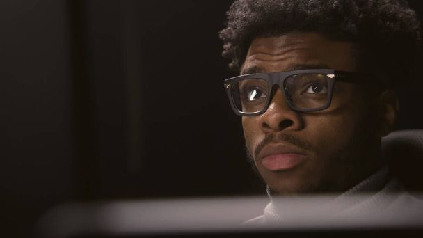Nahaufnahme eines Afroamerikaners, der im Studio mit modernem Design arbeitet oder Filme anschaut. Filmredakteur blickt auf Monitore. Videomaterial spiegelt sich in der Brille wider. Freiberuflichkeit und Fernarbeit. Vergrößern. - Foto, Bild