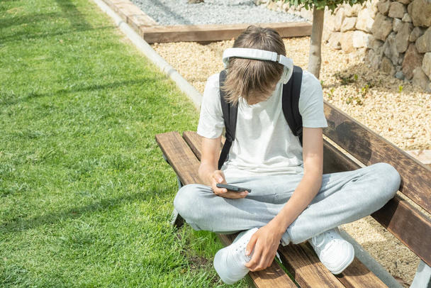 Teenage boy sitting outdoors and using smart phone to listen music with headphones, watching video, writing sms, scrolling news, playing games, chating with friends, using messaging or video chat app. - Foto, immagini