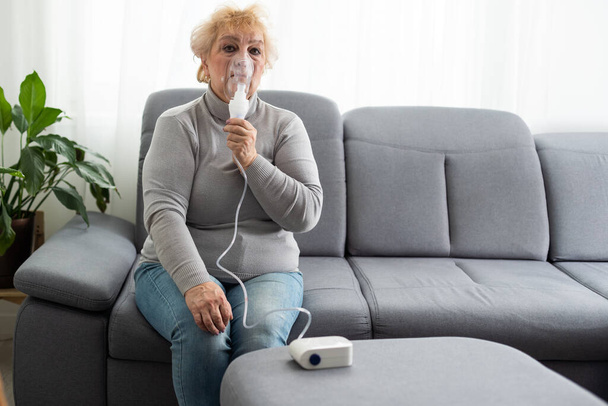 Mujer mayor usando un nebulizador hace inhalación en casa y mira a la cámara. - Foto, imagen
