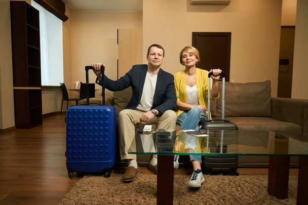 Traveling couple in travel clothes sit in a modern hotel room, they have travel suitcases - 写真・画像