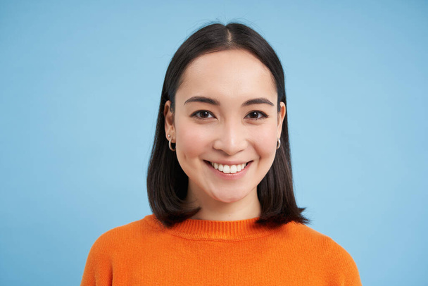 Közelkép portré ázsiai lány tökéletes egészséges mosoly és a természetes szép arc, úgy néz ki, boldog a kamera, áll a kék háttér. - Fotó, kép