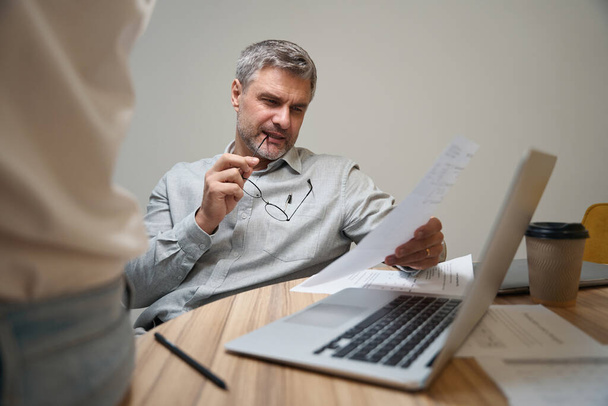 Взрослый мужчина сидит за столом рядом с ноутбуком в коворкинге, держит документ и читает - Фото, изображение