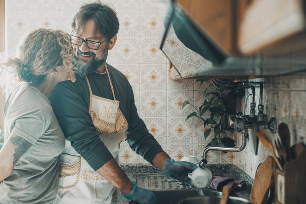 Happy couple adult people in love washing dishes together in the kitchen living at home sharing apartment. Married husband and wife doing housework with smile and happiness. Love lifestyle man woman - Valokuva, kuva
