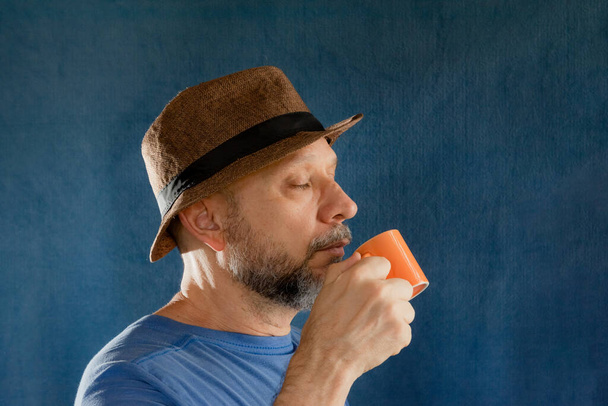 Bearded middle-aged man, wearing a hat, holding an orange colored cup. Isolated on blue background  - 写真・画像