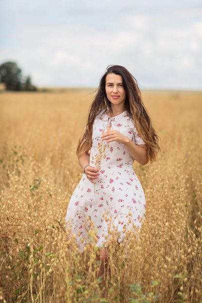 A beautiful brunette woman in a white dress runs along a field of golden chickpeas ear. Stylish girl in the field in windy weather - Фото, изображение
