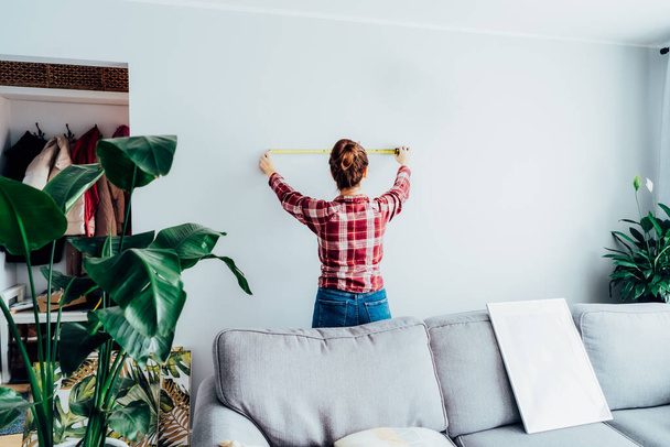 Young woman in plaid shirt doing measuring with a measure tape on the wall. Girl wants to put a picture on the wall at home. Housekeeping work. Doing repair herself. DIY, equality in work concept - Foto, Imagem