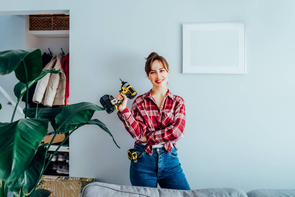 Happy smiling proud of herself young brunette woman in plaid shirt holding electrical screwdriver after she put a picture on the wall at her home. Housekeeping work. Doing repair herself. DIY concept - Foto, immagini