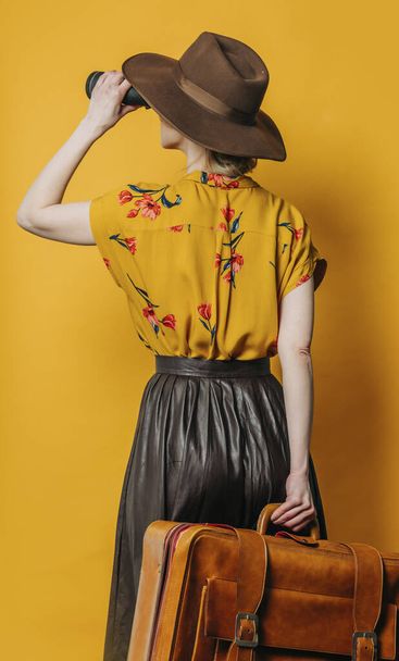 Back side view on stylish blond hair woman in hat and shirt with binoculars and suitcase on yellow background - 写真・画像