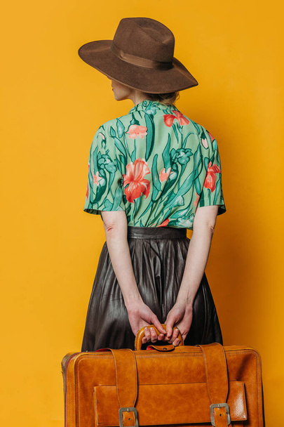 Back side view on stylish blond hair woman in hat and shirt with suitcase on yellow background - Photo, Image
