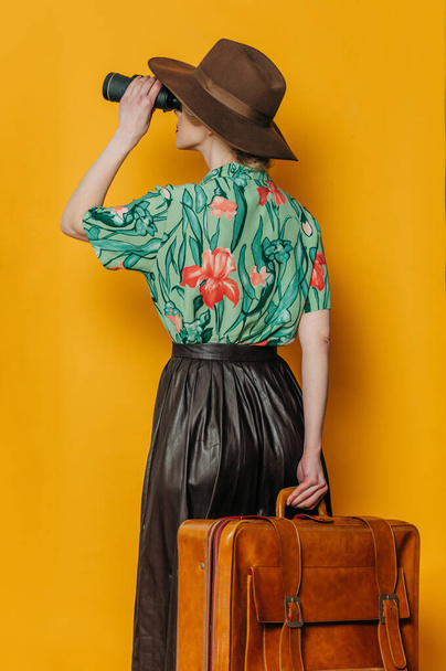 Back side view on stylish blond hair woman in hat and shirt with binoculars and suitcase on yellow background - Photo, Image