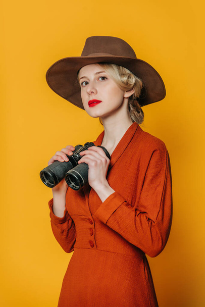 Stylish blond hair woman in hat and brown dress with binoculars on yellow background - Foto, immagini