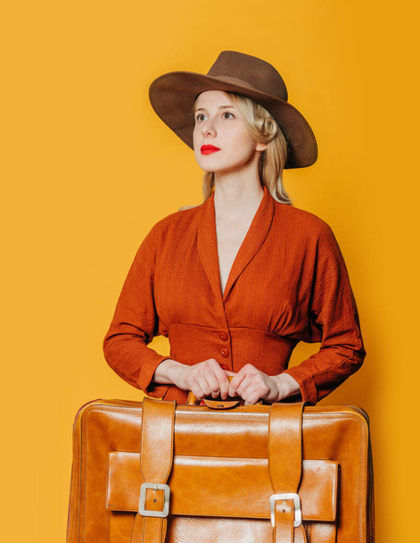Блондинка в винтажной шляпе и коричневом платье с чемоданом на желтом фоне - Фото, изображение