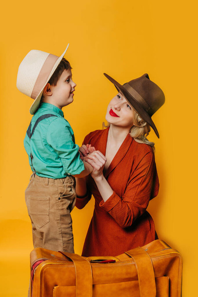 Stylish mother and son in vintage clothes with suitcase on yellow background - Foto, Imagem