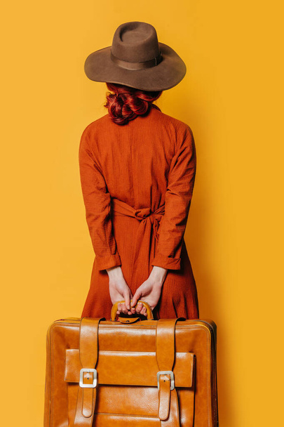 Back side view on stylish redheaded woman in hat and brown dress with suitcase on yellow background - 写真・画像