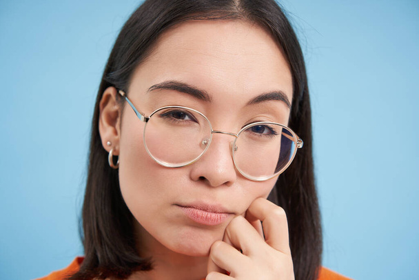 Nahaufnahme Porträt einer asiatischen Frau sieht fasziniert aus, trägt eine Brille, schielt nachdenklich, denkt nach, nimmt an, steht vor blauem Hintergrund. - Foto, Bild