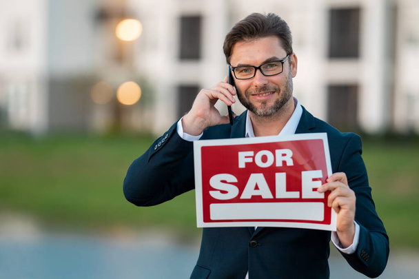 Successful real estate agent in a suit holding for sale sign near new apartment. Real estate agent with home loan contract, selling home. Realtor or real estate agent shows board for sale - Foto, Imagem