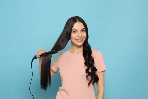Beautiful happy woman using hair iron on light blue background - Foto, afbeelding