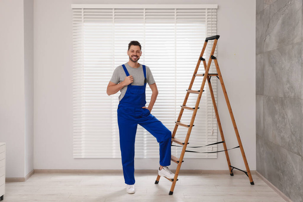 Worker in uniform and stepladder near horizontal window blinds indoors - Foto, imagen