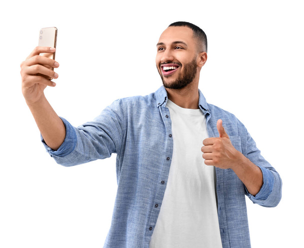 Uśmiechnięty młody człowiek robi sobie selfie za pomocą smartfona i pokazuje kciuki na białym tle - Zdjęcie, obraz