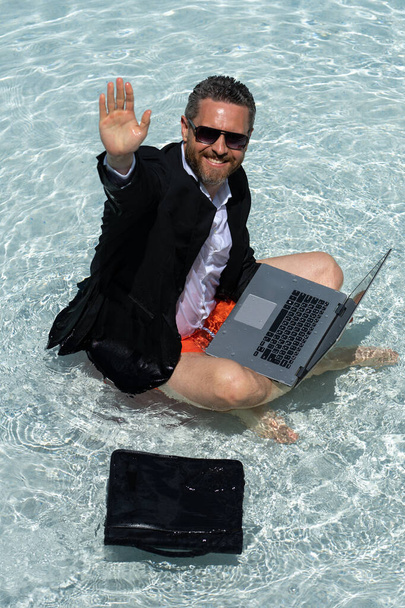 Crazy business man in a business suit works for a laptop standing in the water in pool. Remote work. Crazy freelancer. Business and summer. Business man working online with computer laptop in water - Фото, изображение