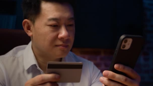 Χαμογελώντας χαρούμενος Ασιάτης στο σκοτεινό βράδυ στο σπίτι γραφείο με κινητό τηλέφωνο χρησιμοποιώντας πιστωτική κάρτα Κορεάτης επιχειρηματίας κάνουν την κράτηση πληρωμής ανέμελη με εύκολη online έκπτωση κινητό τηλέφωνο ψώνια - Πλάνα, βίντεο